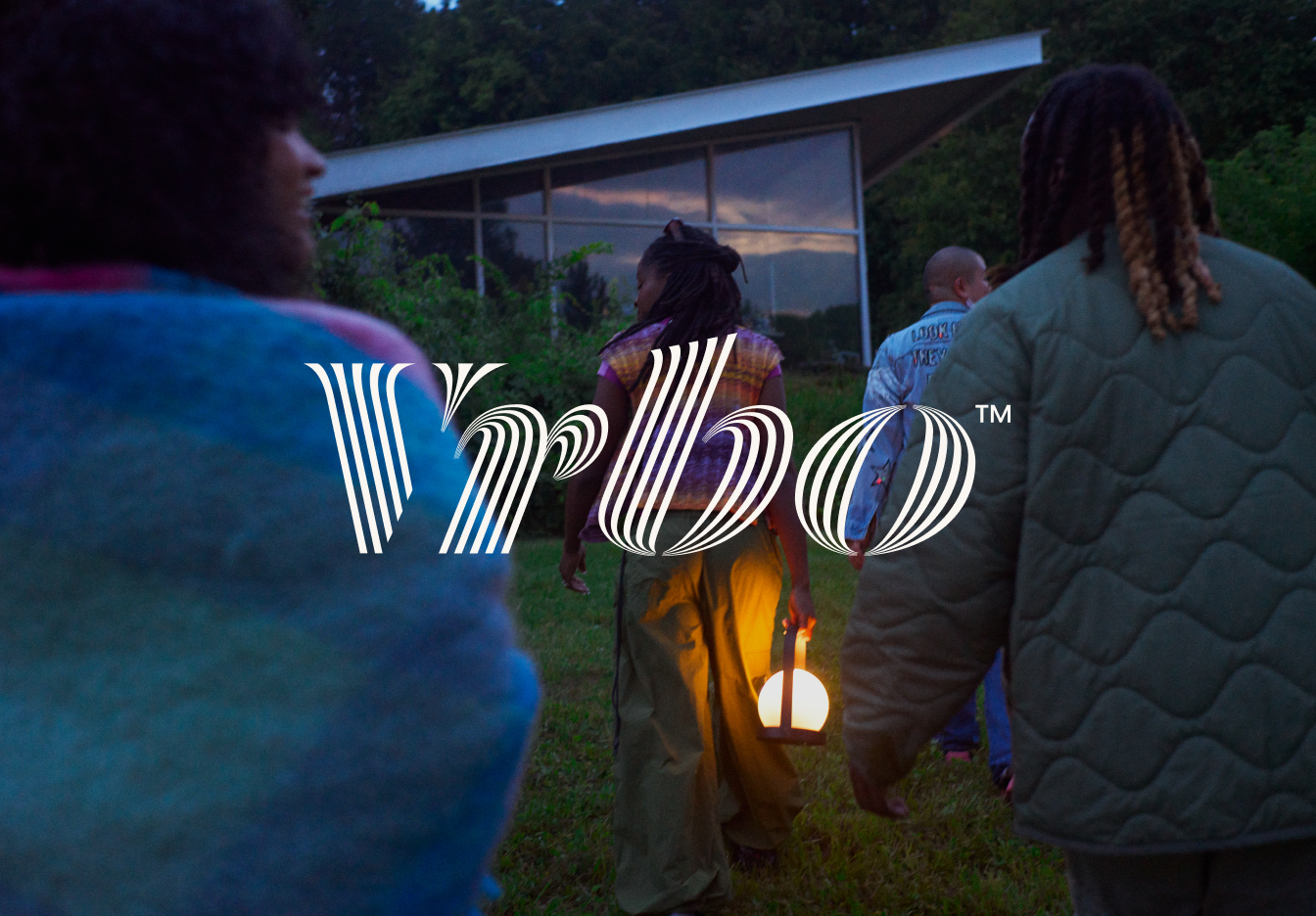 Un gruppo di persone cammina in direzione di una casa e una di loro ha una lanterna in mano. Sopra l'immagine è applicato il logo Vrbo.