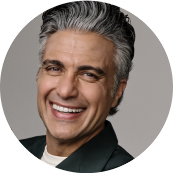 Jaime Camil smiling, wearing a dark green jacket, in front of a neutral background.