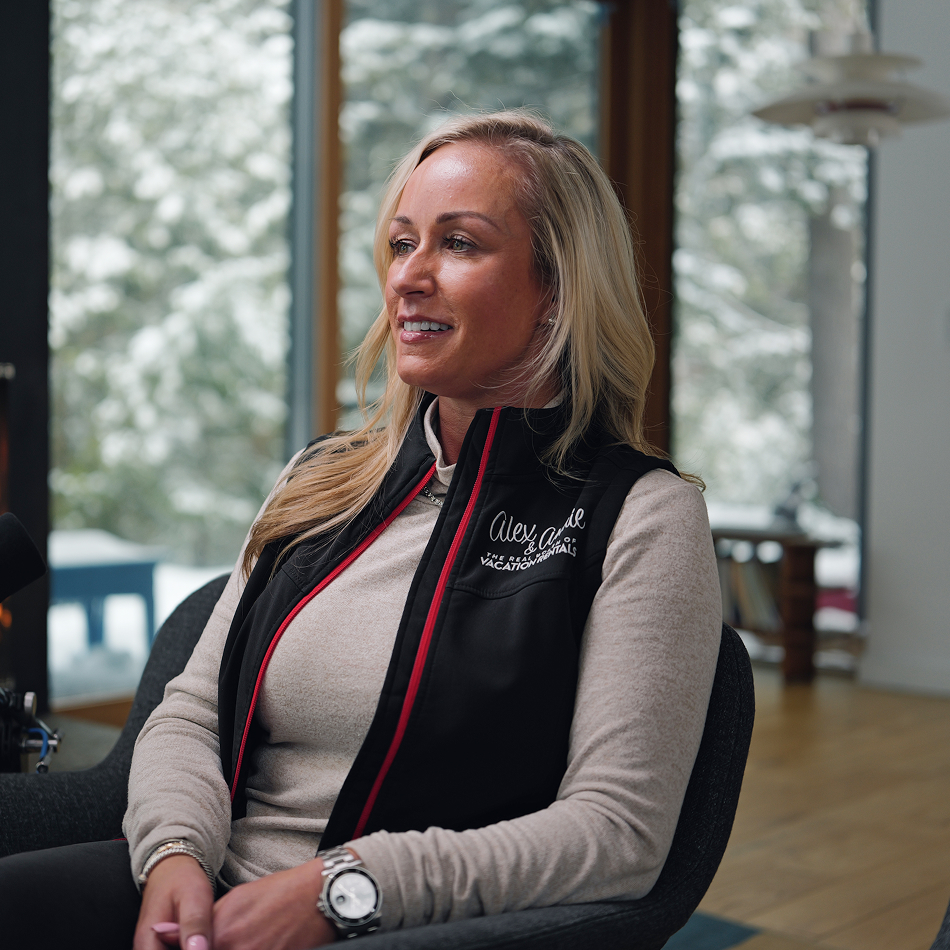 Alex Husner seated in a chair during a podcast-style recording, wearing a black vest over a light long-sleeve top, with snowy trees visible through large windows in the background.