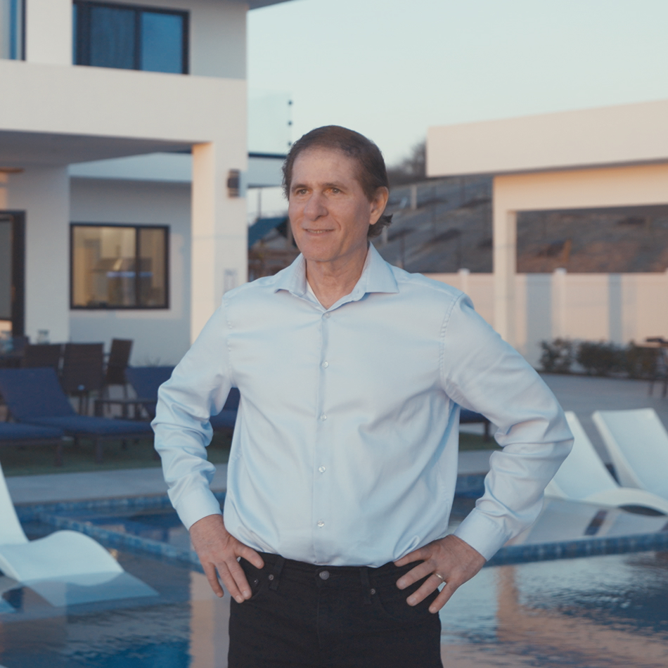 Alex Lluch in a light blue dress shirt standing by a modern swimming pool with his hands on his hips, with a contemporary house and patio furniture in the background.