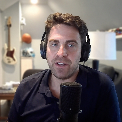 Brandon Ehrhardt wearing headphones speaks into a studio microphone in a warmly lit home recording setup, with a guitar and framed photos softly blurred in the background.