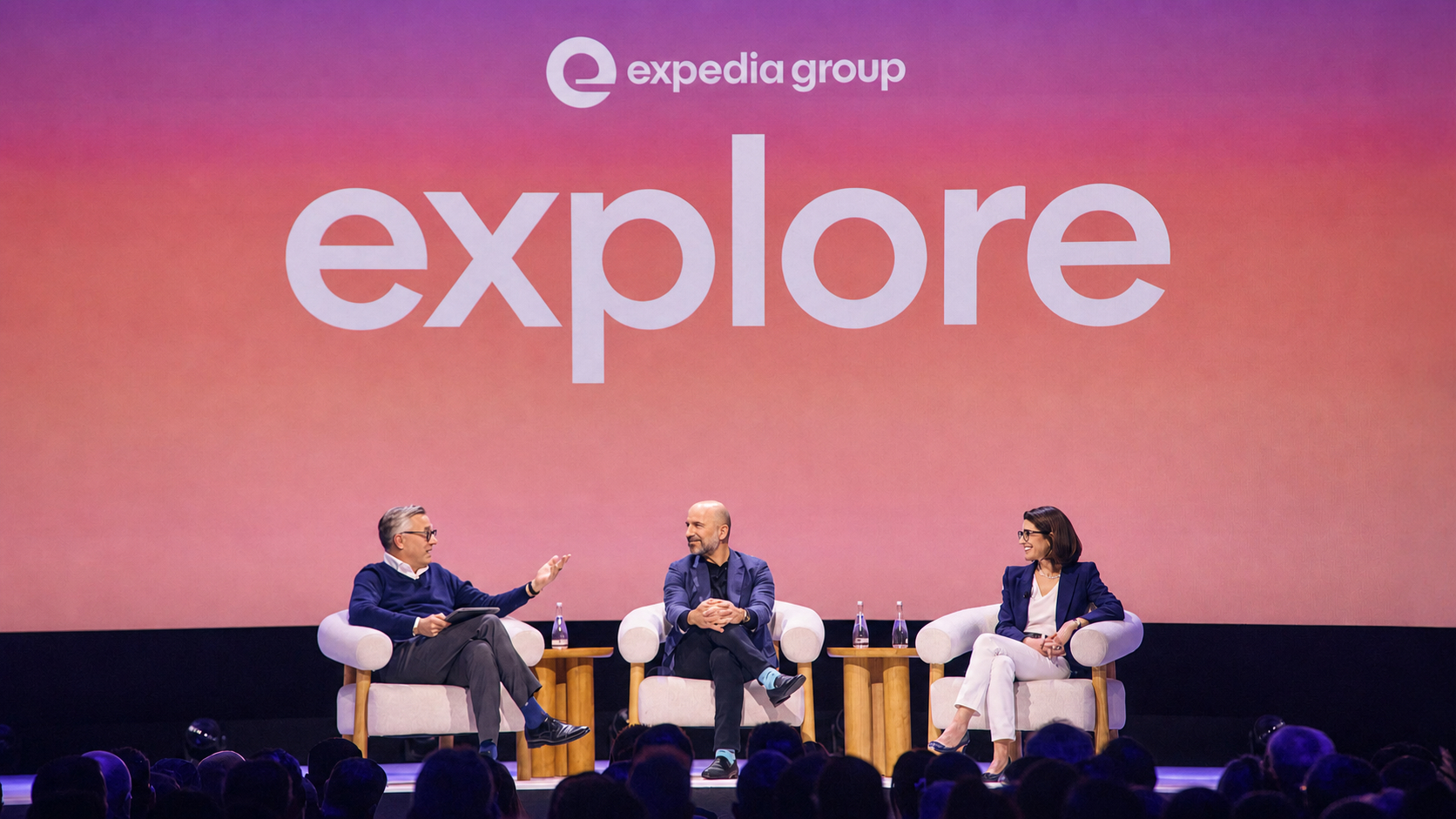 Alfonso Paredes, Dara Khosrowshahi, and Ariane Gorin seated onstage in white armchairs during a panel discussion, with small round tables between them and an audience visible in the foreground. A large pink-to-orange gradient screen behind them displays the Expedia Group logo and the word “explore” in oversized white letters, framing the session as a branded conference conversation.