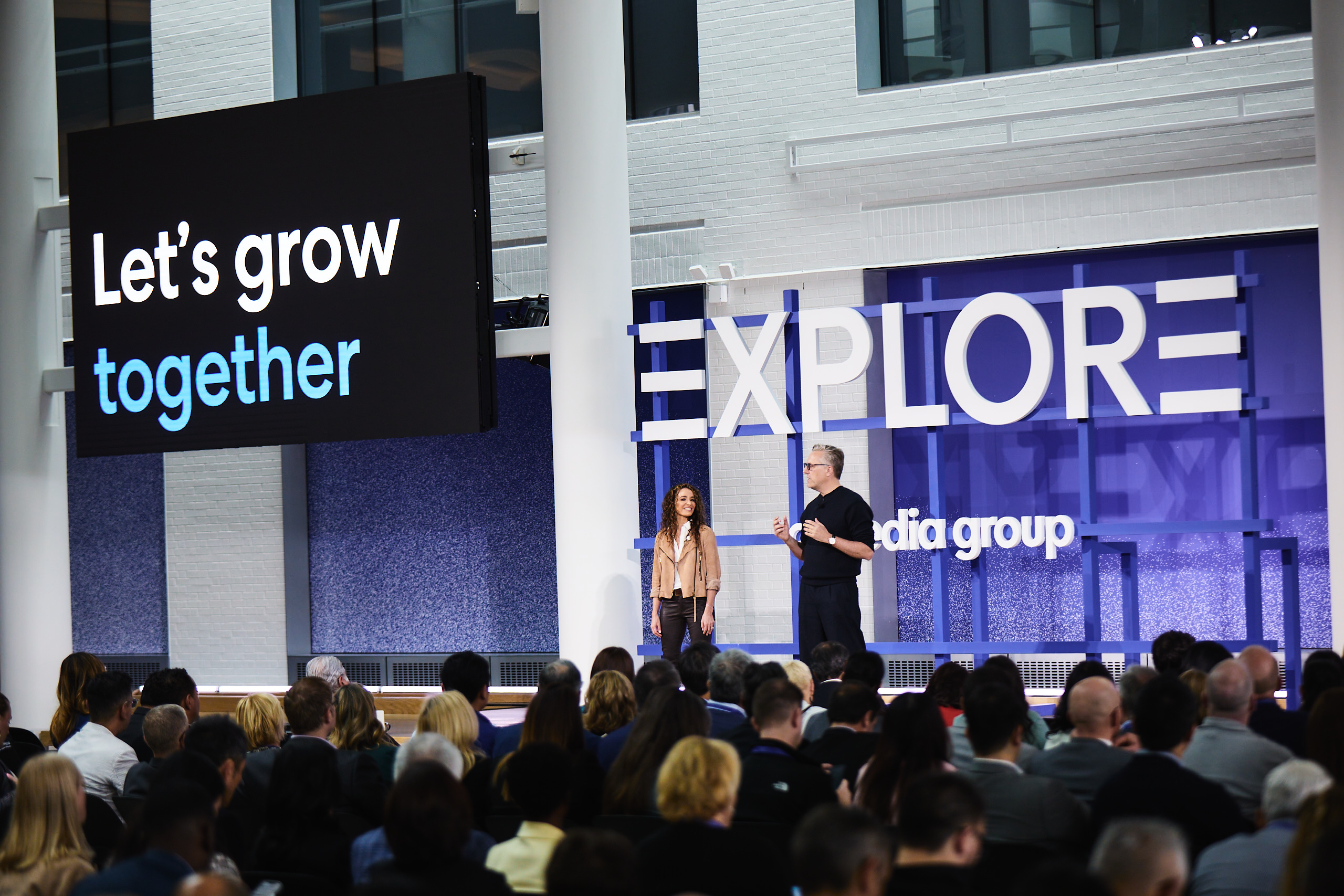 A woman and a man stand on a stage in front of a crowd and large sign reading "EXPLORE" and screen reading "Let's grow together."