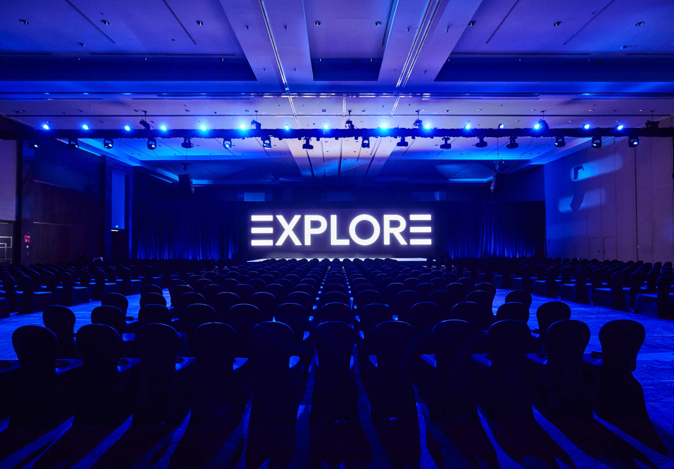 Large conference hall with rows of empty chairs facing a stage with a bright “EXPLORE” sign illuminated in blue lighting.