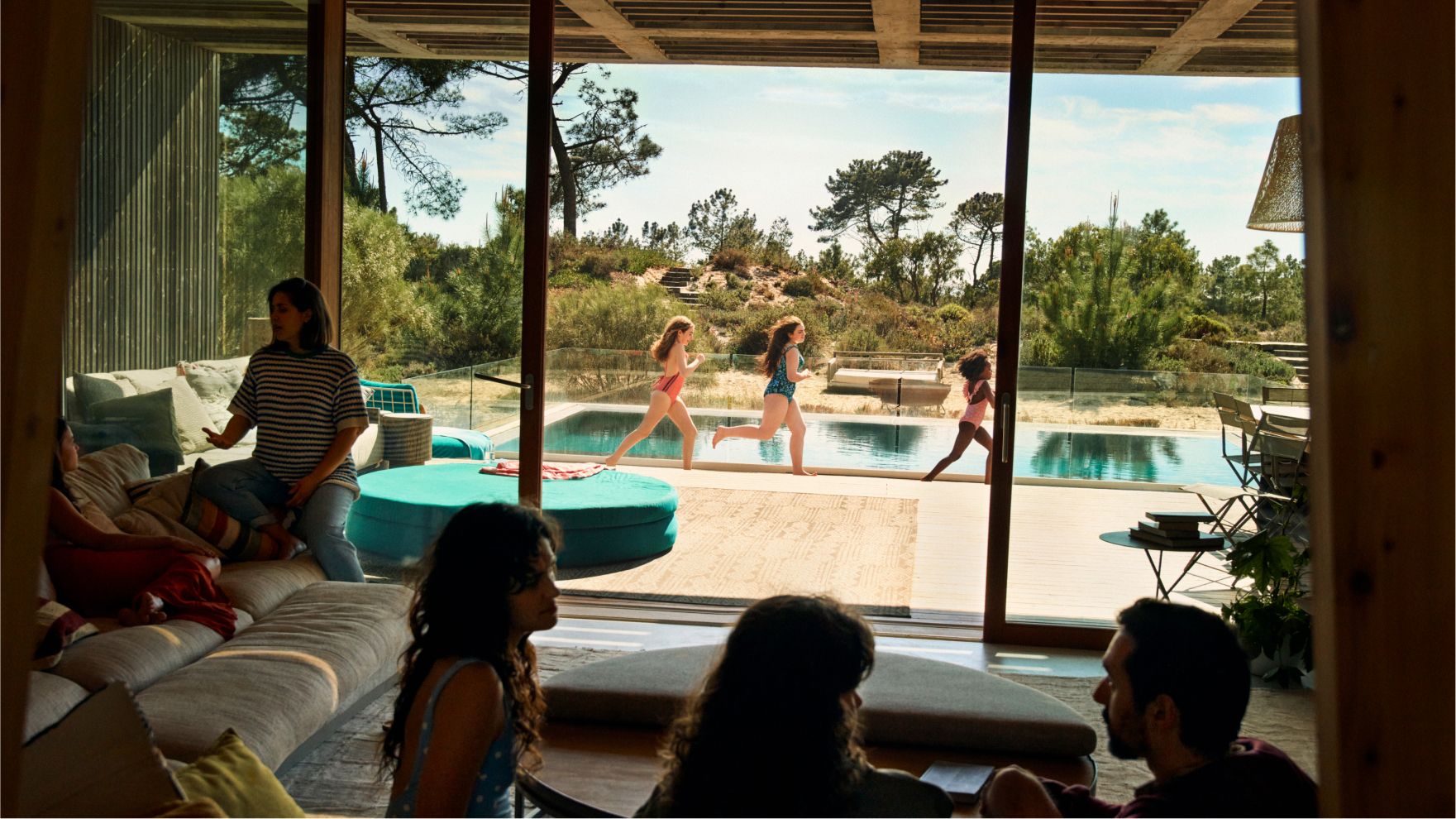 Children in swimsuits run beside an outdoor pool while family members gather and talk inside a modern vacation home with large windows
