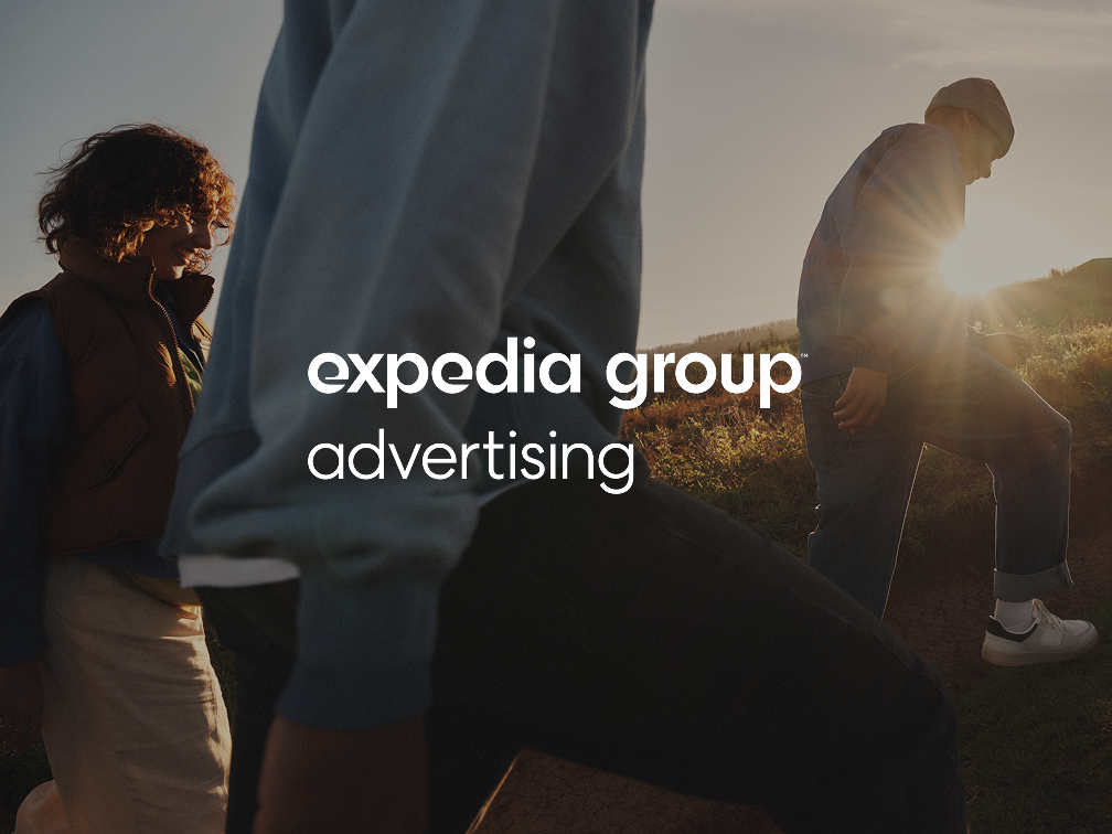 A group of three friends dressed in warm clothing are hiking up a hill in the countryside while the sun sets behind them. Rays of the sunlight shine just over the horizon creating a majestic glare, silhouetting one of the climbers as they ascend the hill.
