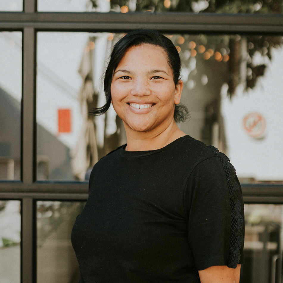 Portrait of Helen Melluish smiling warmly, wearing a black short-sleeve top, standing in front of a large glass-paneled storefront window with string lights visible inside.
