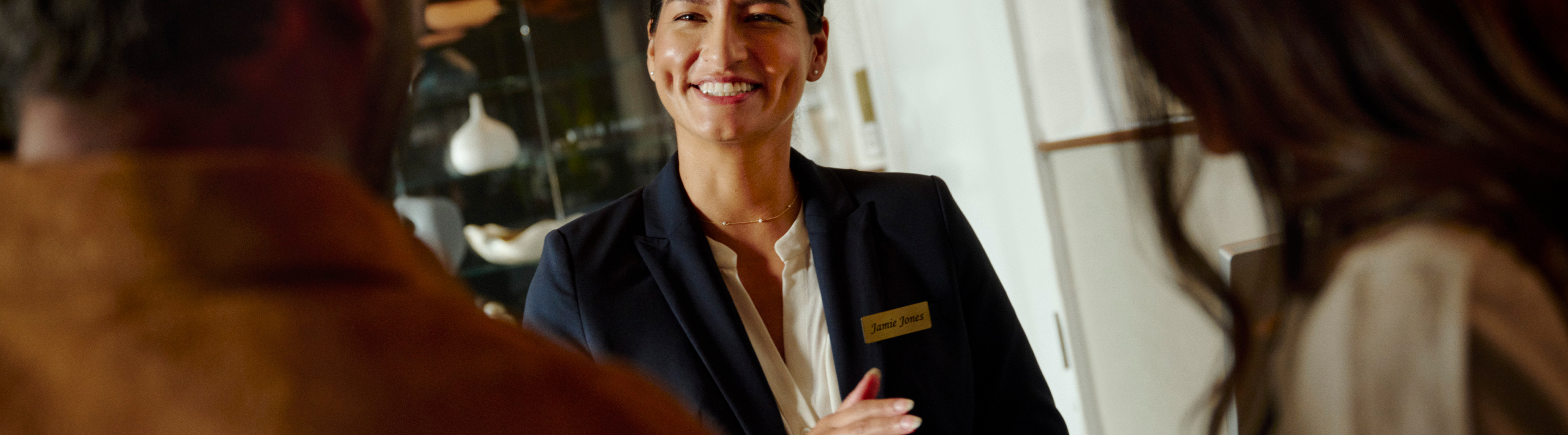 A smiling hotel concierge in a navy blazer with a name badge warmly greeting guests at the front desk