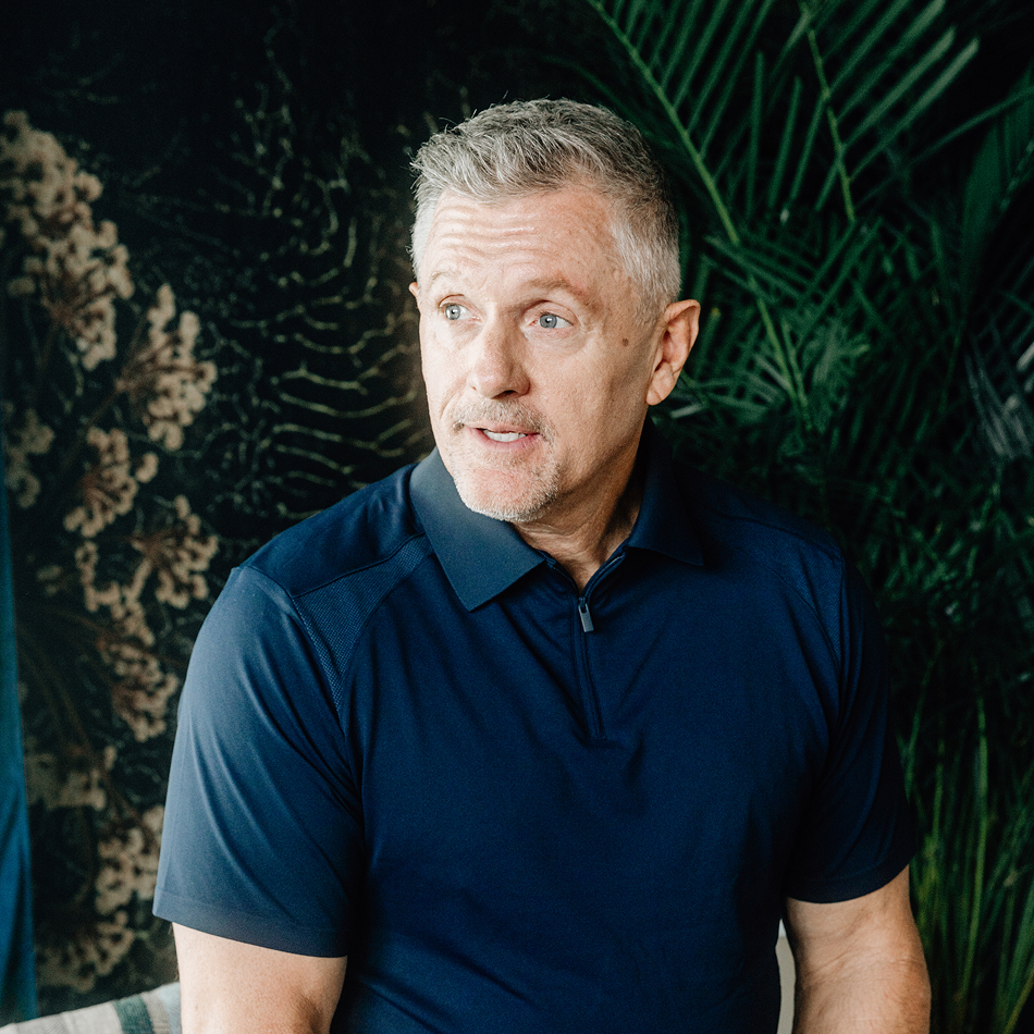 Portrait of Kyle Whittingham with short gray hair wearing a navy zip-collar polo shirt, looking off-camera with a relaxed expression against a lush backdrop of tropical palm leaves and botanical wallpaper.
