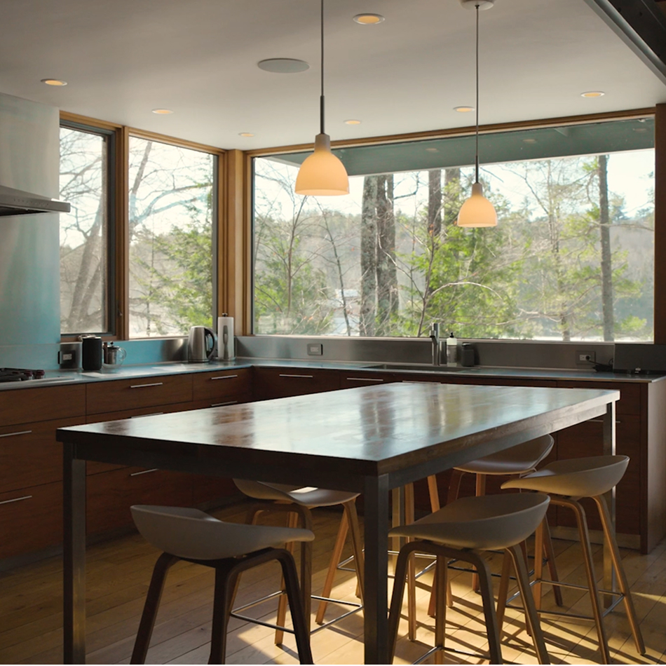 A modern kitchen with a dark wood island, tall stools, pendant lights, and wraparound windows looking out onto a wooded landscape.