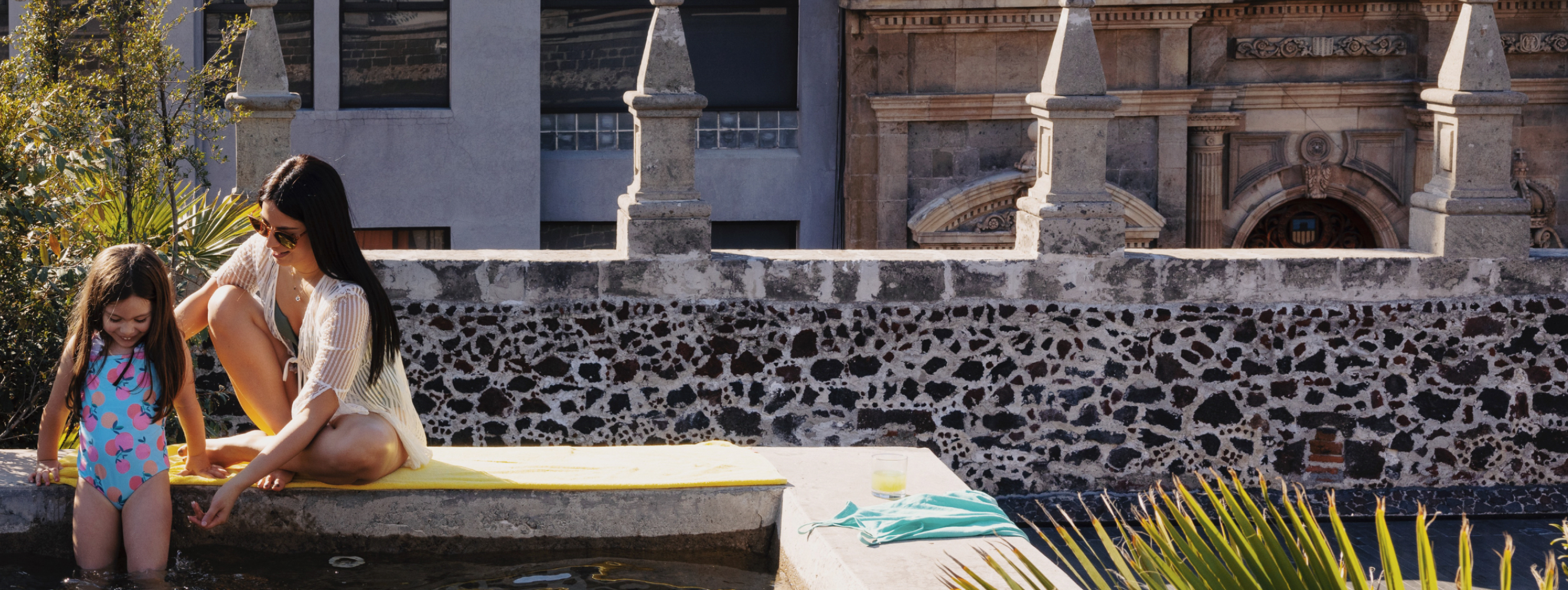 A mother and daughter sit at the edge of a courtyard pool, dipping their feet in the water together.