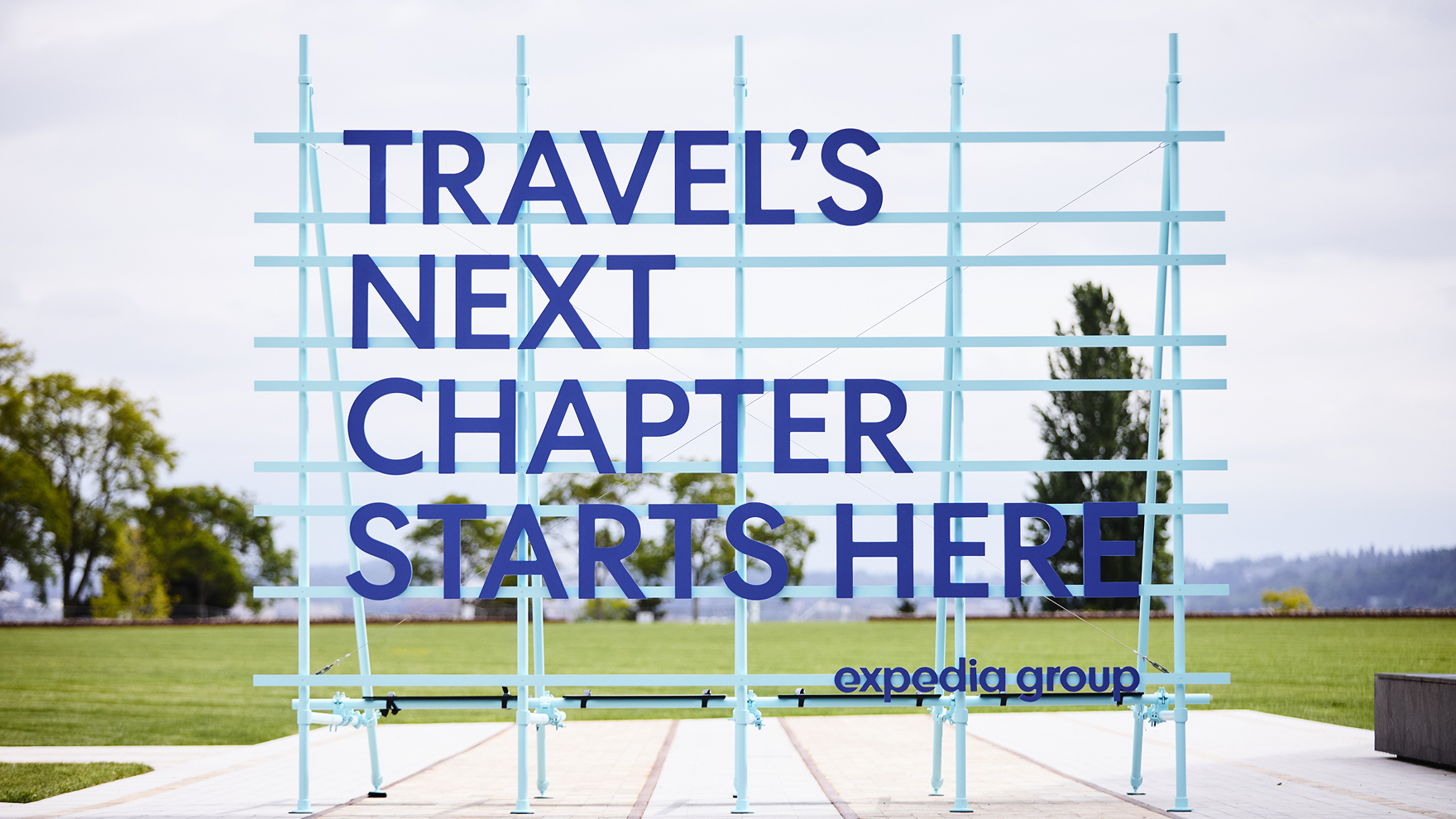 Large outdoor sign reading “Travel’s Next Chapter Starts Here” with Expedia Group logo, placed in a landscaped area.