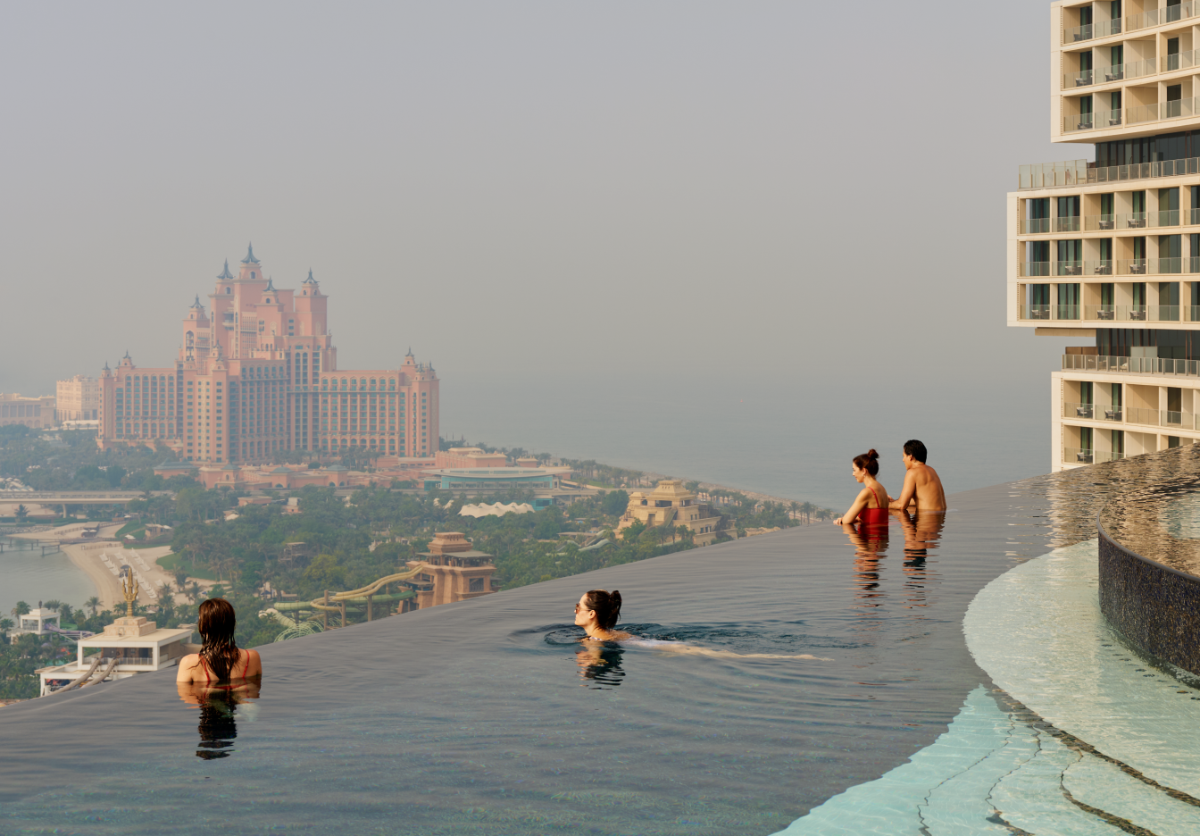  Group of hotel guests swimming in a roof-top infinity-edge pool at a luxury hotel overlooking Dubai.