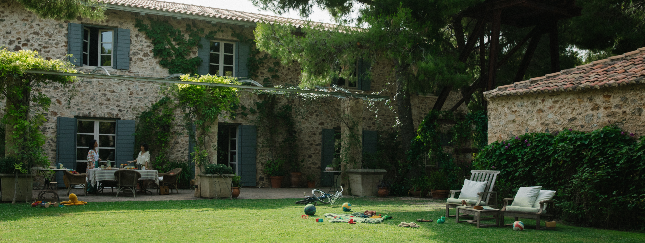 Lush backyard of a brick-walled home in Athens, Greece with children's toys lying on the grass and two women talking by an outdoor table.