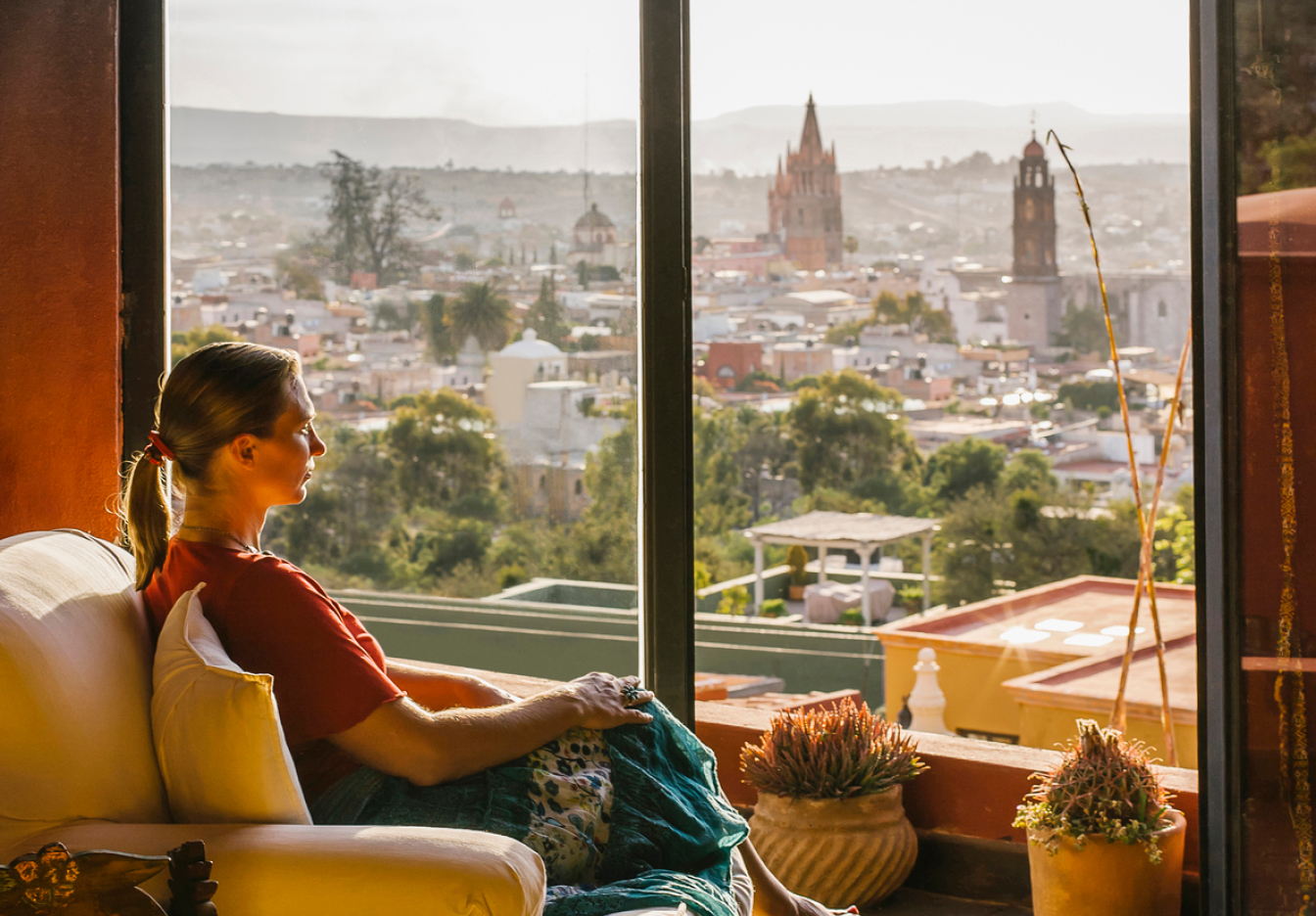 A woman in a red shirt sits in an armchair beside a large window, looking out over a sunlit city with colorful rooftops, trees, and prominent church towers.