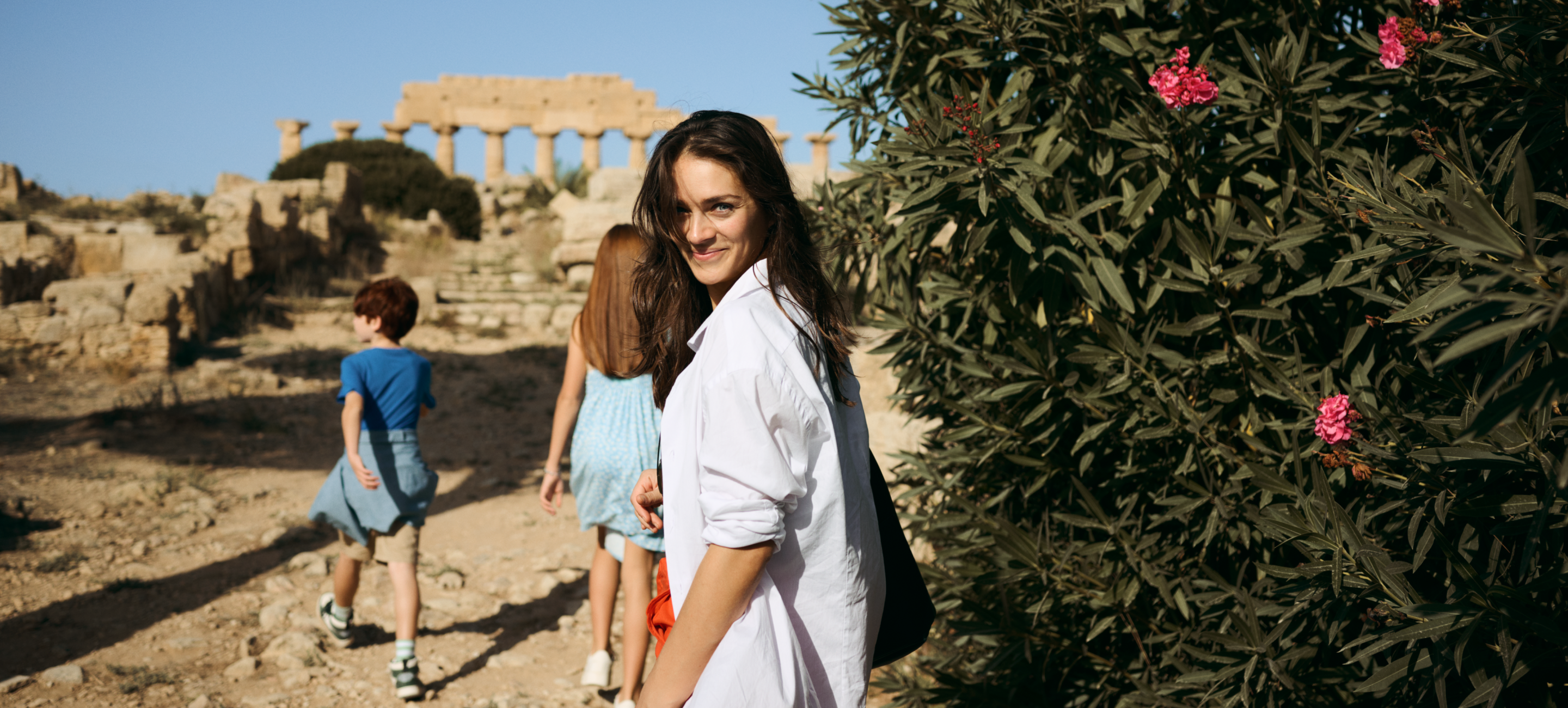 Woman looking over her shoulder and smiling with two children and rocky palatial ruins in the background.