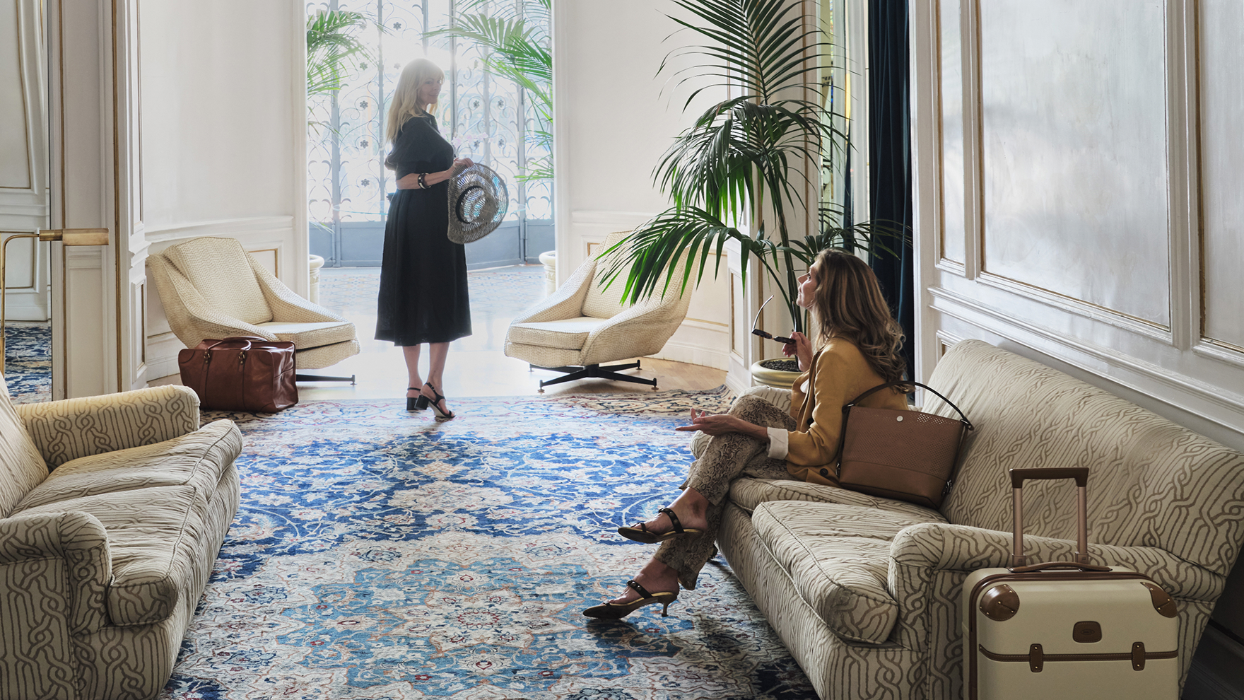 Two women relax in an elegant hotel lobby with luggage nearby; one stands near the bright entry holding a hat, while the other sits on a couch with sunglasses in hand.