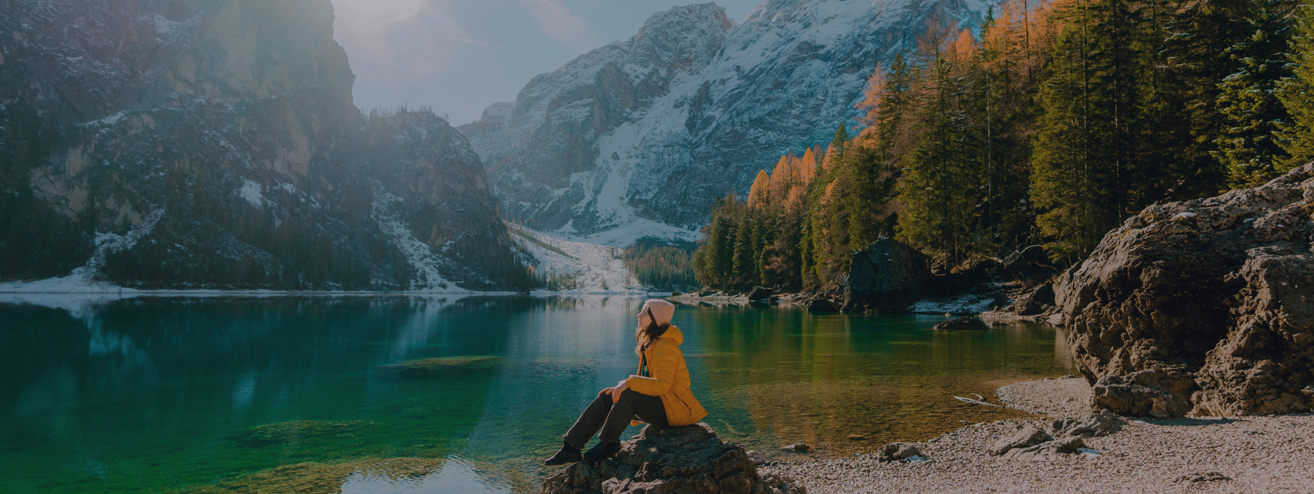 Person in yellow jacket sitting by a clear mountain lake surrounded by pine forest and snow-covered peaks