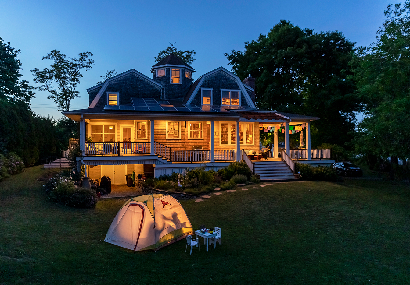 Twilight view of a large lit-up vacation home with solar panels and wraparound porch, with a glowing camping tent and small table with chairs set up on the front lawn.