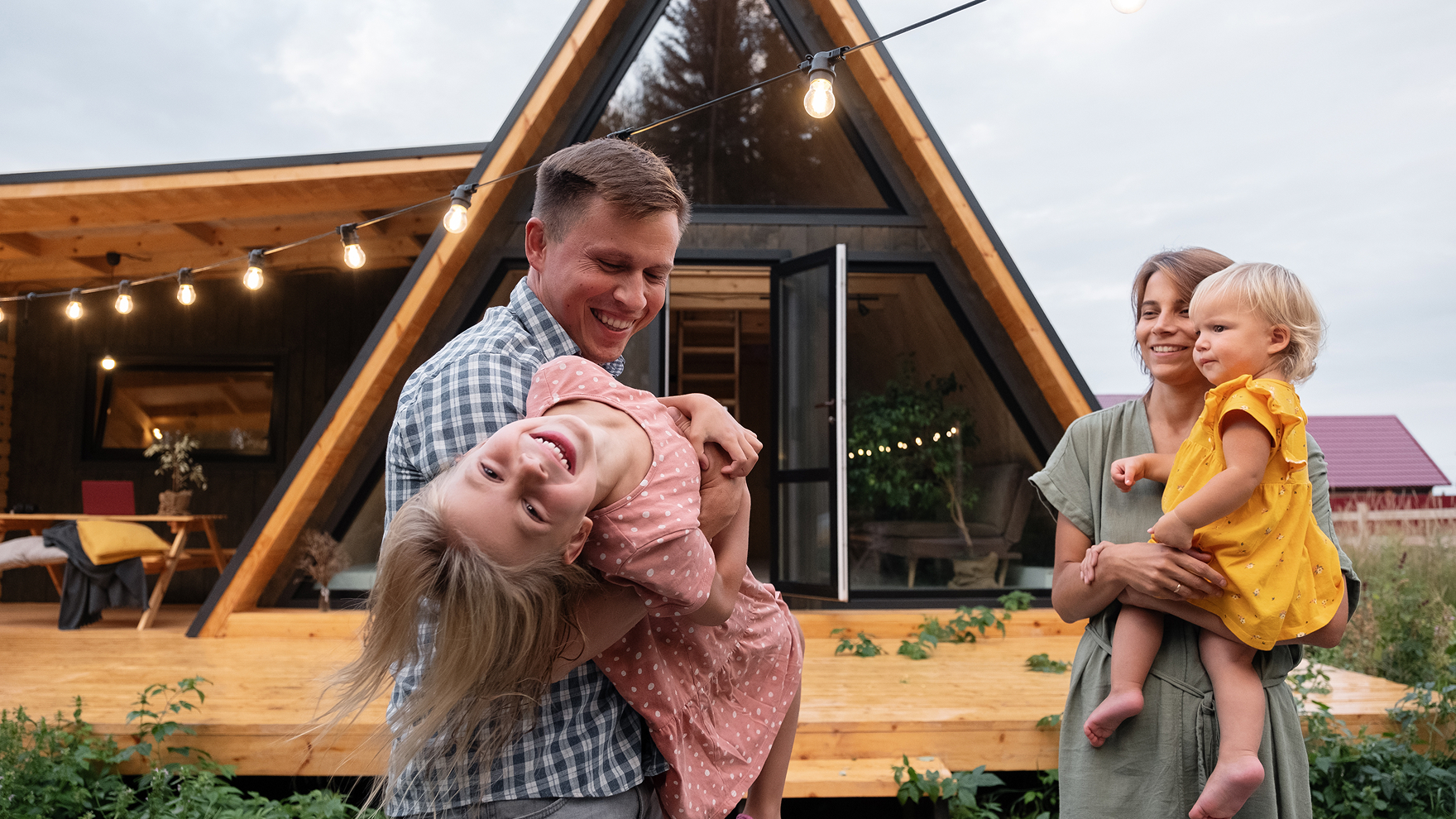 A smiling family stands outdoors on the wooden porch of a modern A-frame cabin decorated with string lights. A man holds a laughing little girl, upside down in his arms, while a woman hugs a blonde toddler in a yellow dress. The background features grass and a cozy cabin, creating a warm, inviting atmosphere