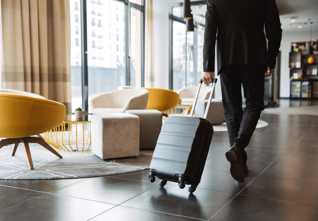 Person in business attire walking through a modern hotel lobby pulling a rolling suitcase.