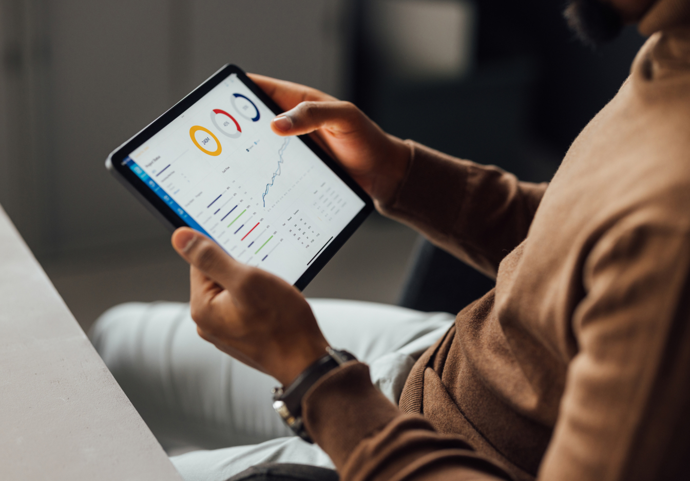 Person sitting indoors, holding a tablet displaying colorful data charts and analytics, with focus on the screen and hands.
