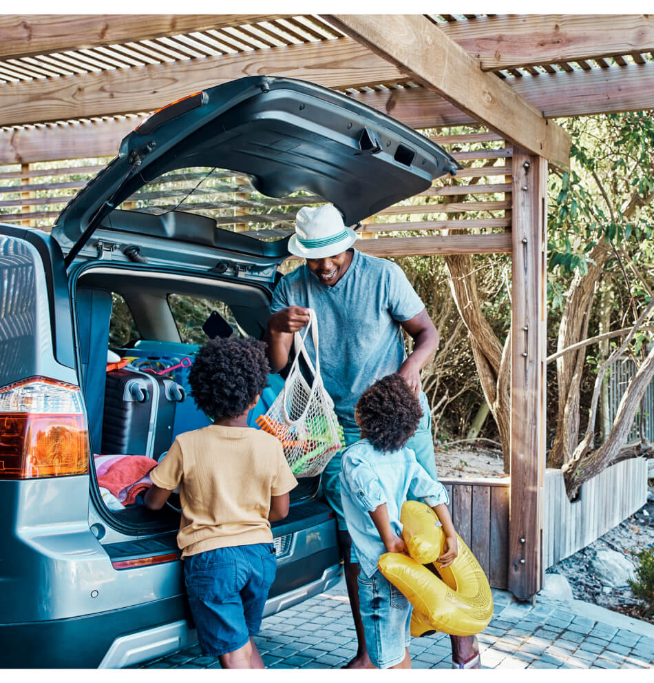 A father loading luggage into the trunk of their car while two children watch.