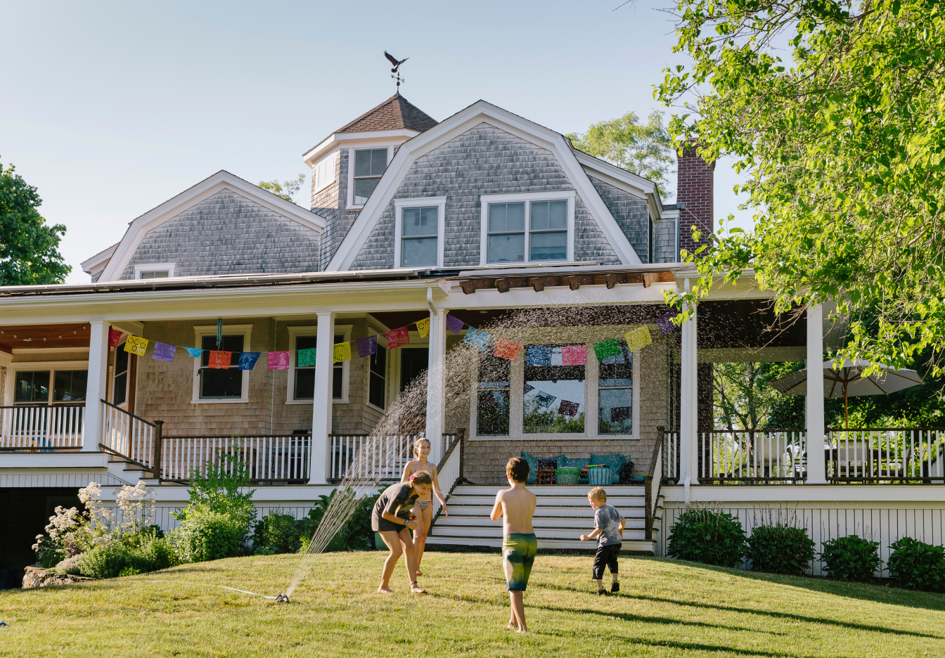 Three children play in the spray of a lawn sprinkler on a sunlit grassy yard in front of a large gray shingle-style house with a wraparound porch, white columns, colorful stringed flags, and broad front steps leading up to the entrance.