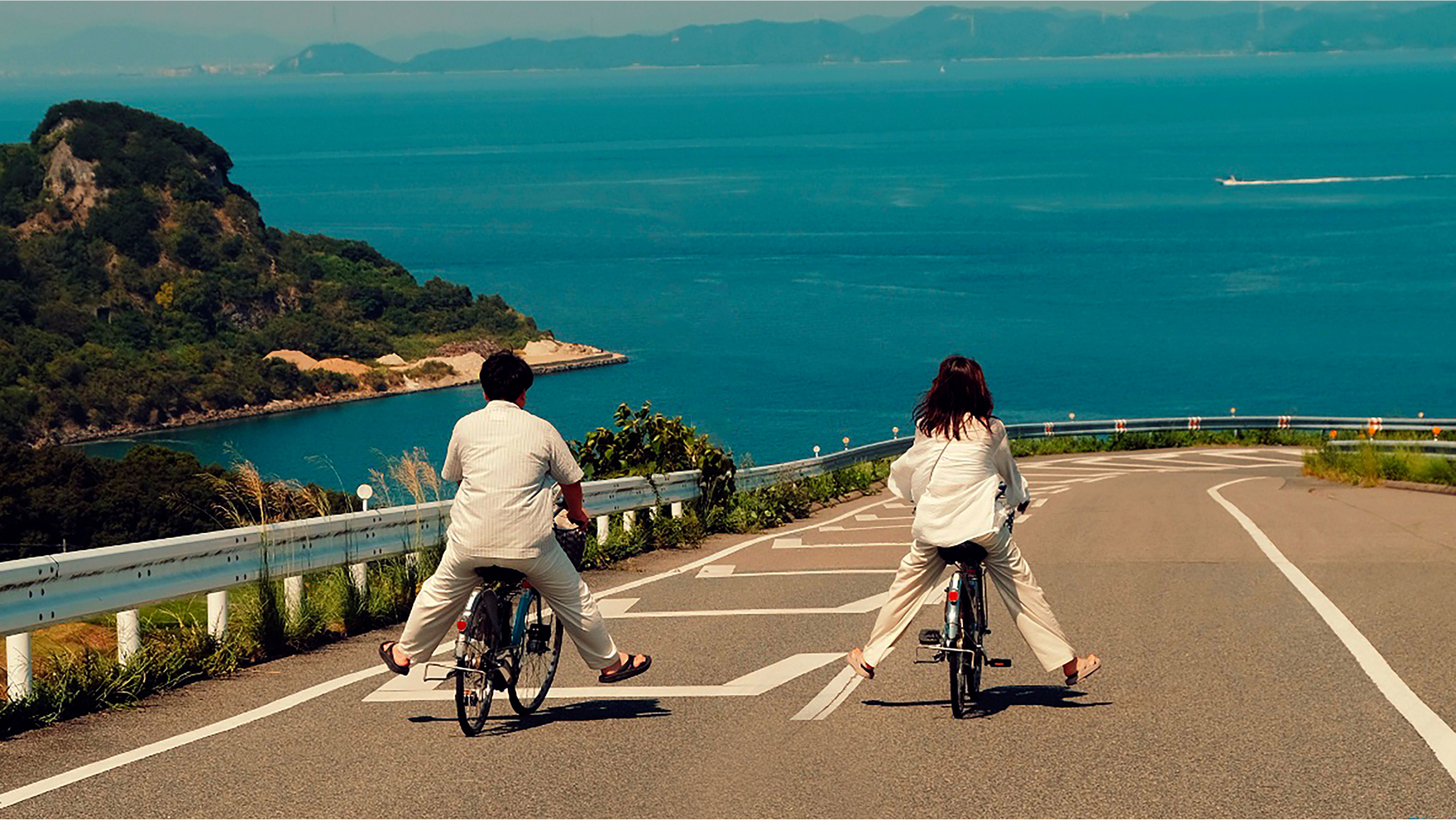 Two people riding bicycles downhill on a coastal road with the ocean and distant islands in the background.
