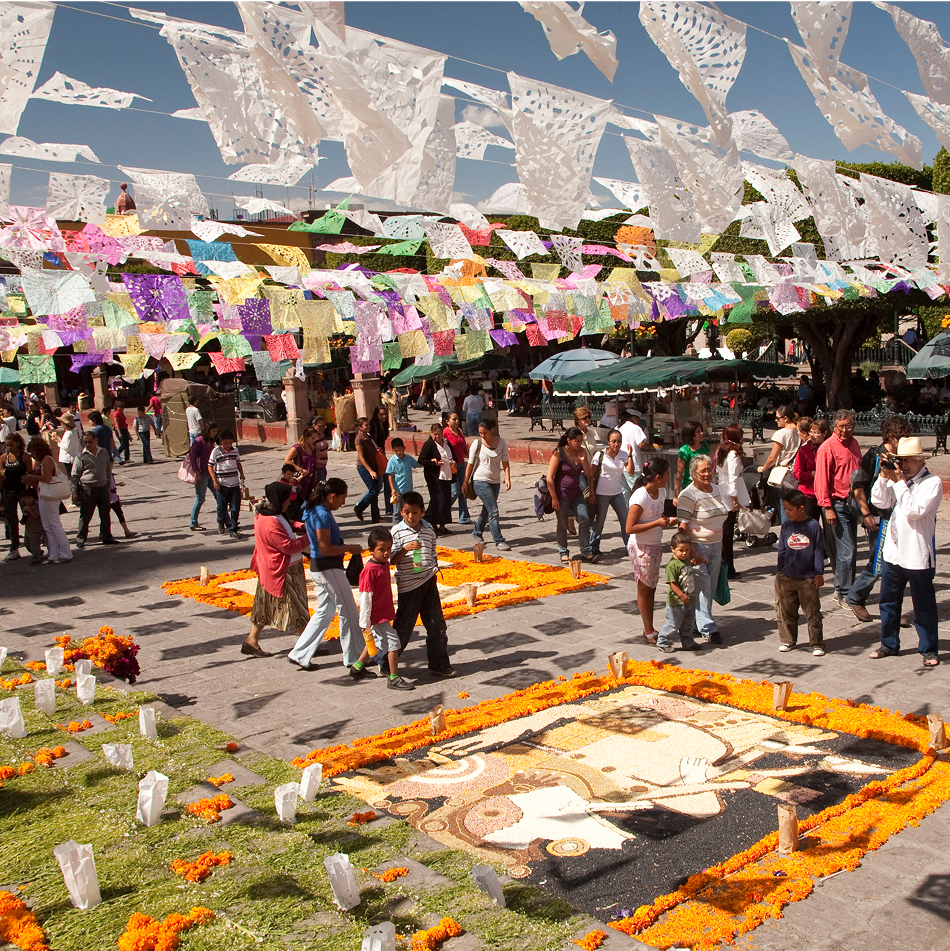 Crowd of people walking through a decorated plaza with colorful papel picado banners and marigold-bordered street art for Day of the Dead celebrations.