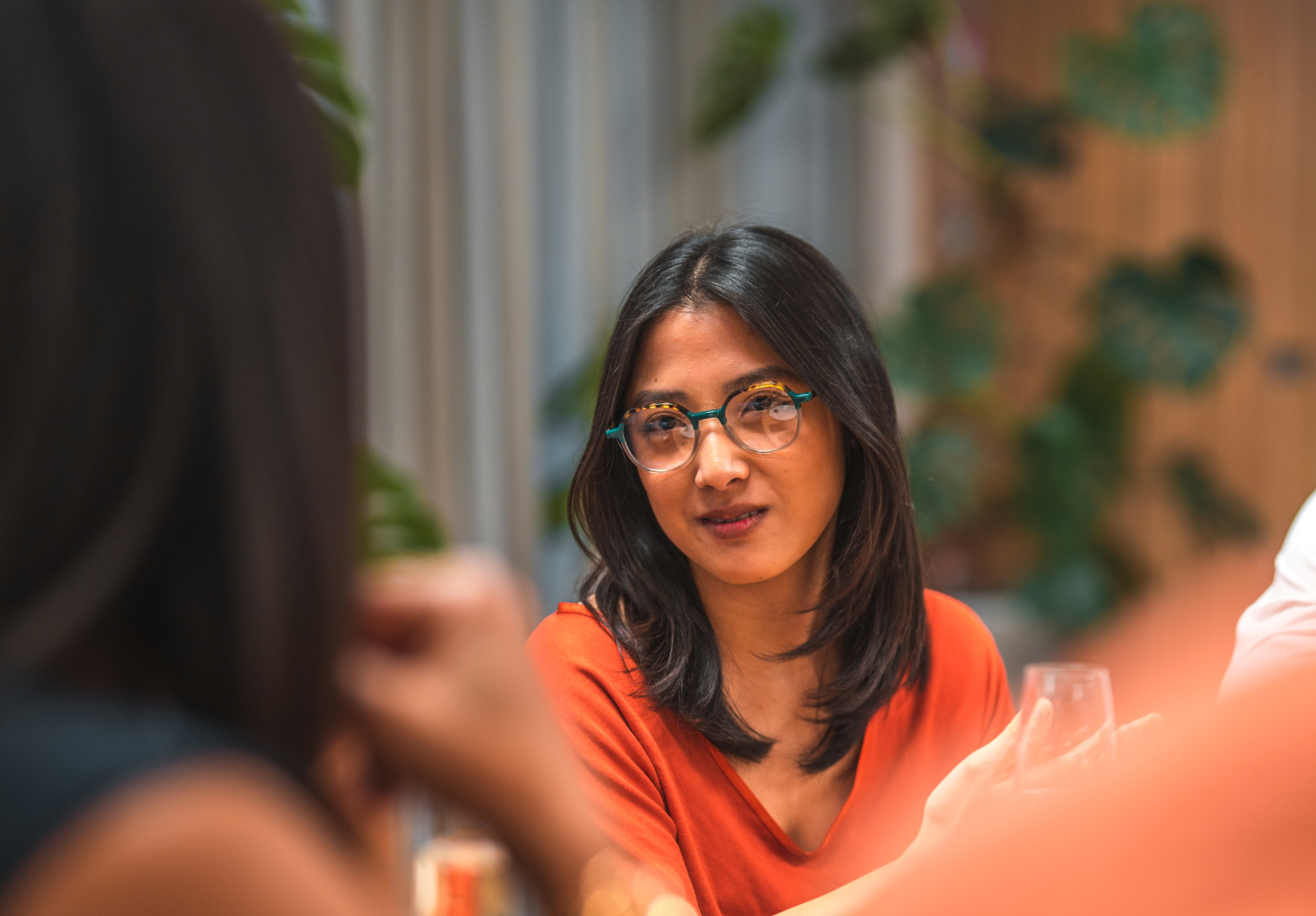 Person in an orange top wearing glasses, listening during a conversation at a dinner table indoors.