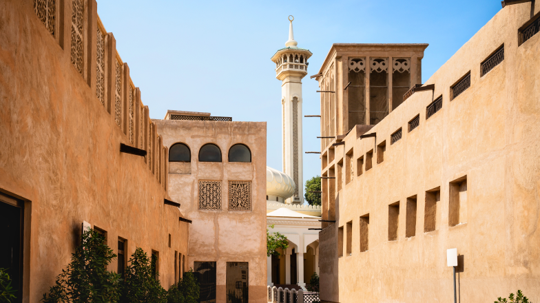 A traditional Middle Eastern alleyway in Old Dubai featuring beige-colored historic buildings and a mosque tower in the background.