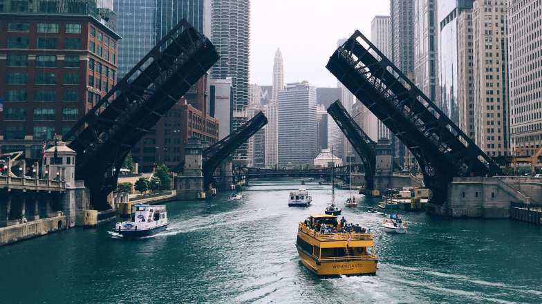 A daytime view of the Chicago River with multiple drawbridges raised to allow boats to pass. Tall skyscrapers line the river, creating an urban skyline.