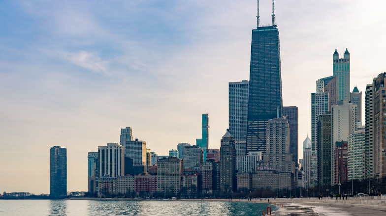 A sunset view of the Chicago skyline from the edge of Lake Michigan, featuring the iconic John Hancock Center and surrounding high-rise buildings.