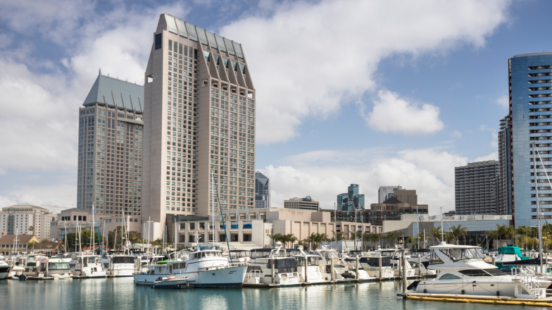 A marina in San Diego, California, with luxurious yachts and boats docked in calm blue waters. In the background are modern high-rise buildings, including the distinctive Manchester Grand Hyatt.