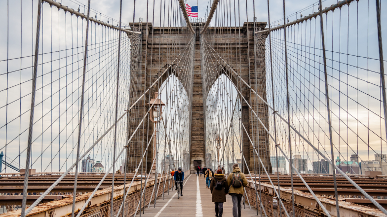 Groups of pedestrians crossing the Brooklynn Bridge with a view of sky scrapers in the background.