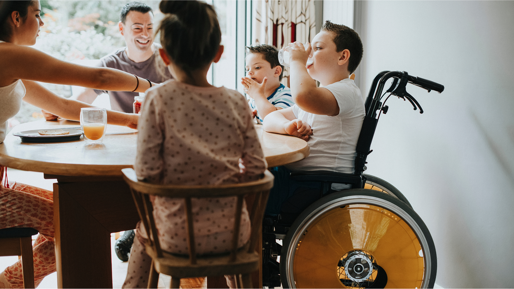 Family enjoying breakfast together in a bright kitchen with a child on a wheelchair beside them, suggesting an accessible home environment.
