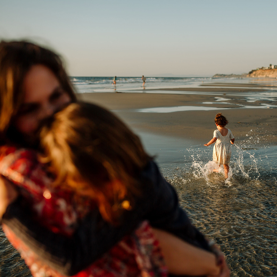Child in a white dress splashing through shallow water on a beach while two people embrace in the foreground