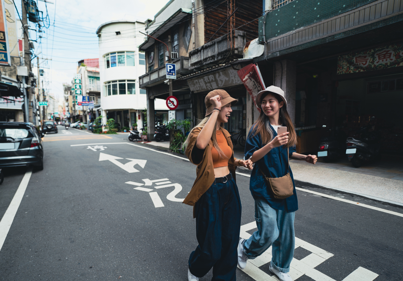 Two young women laughing and walking across a street in an Asian city, one wearing a cap and orange top, the other in a denim jacket and white hat holding a phone, with local shopfronts and signage in the background.