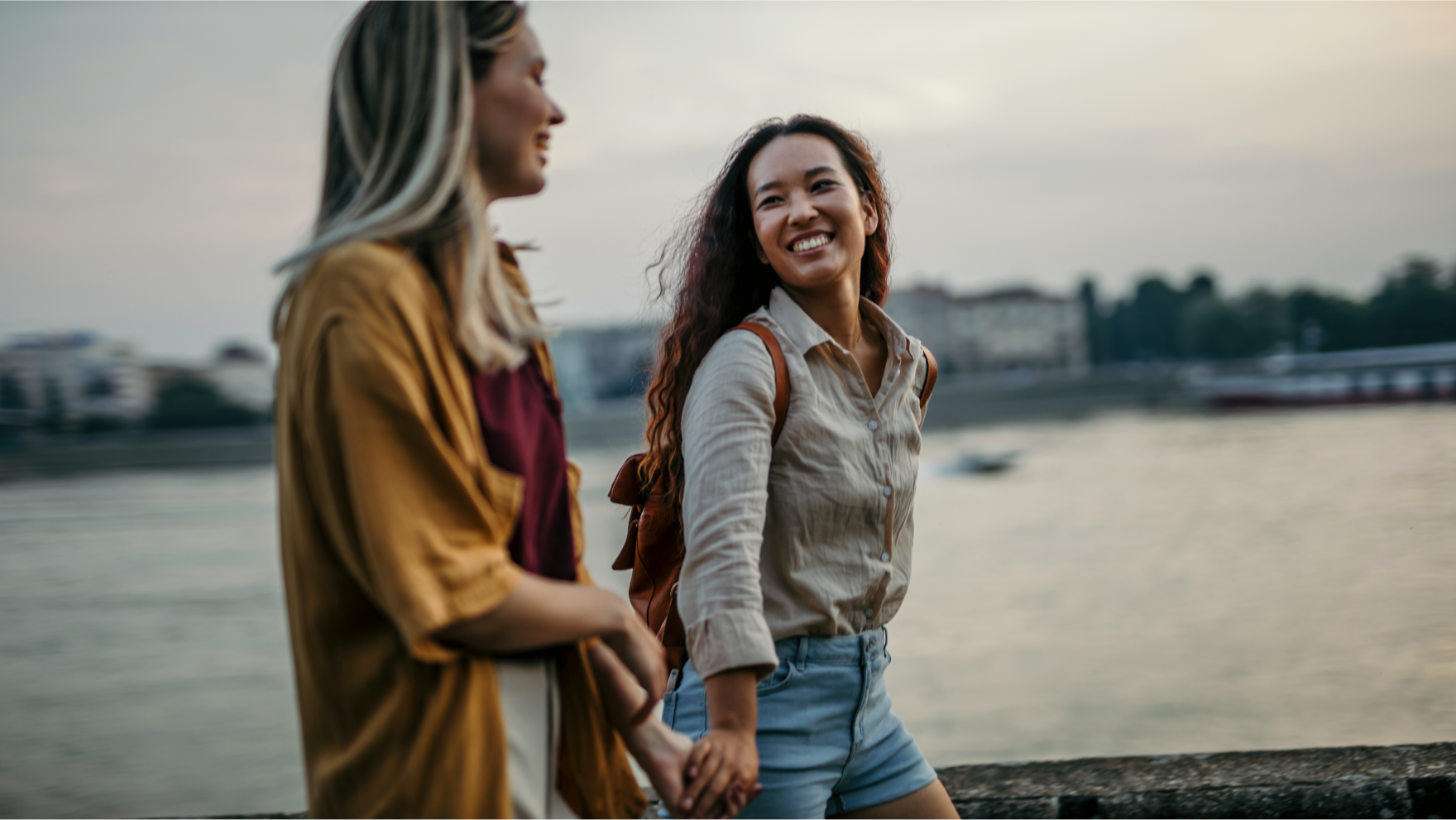 A female couple holding hands while strolling along a riverwalk at dusk.