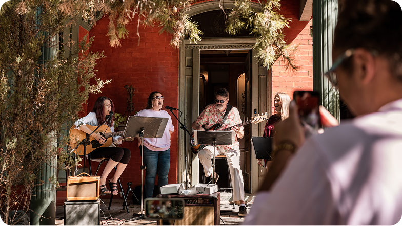A small band performing live outside a red brick building, featuring two guitarists, two singers, and a bass player, while a person in the foreground captures the moment on a smartphone.
