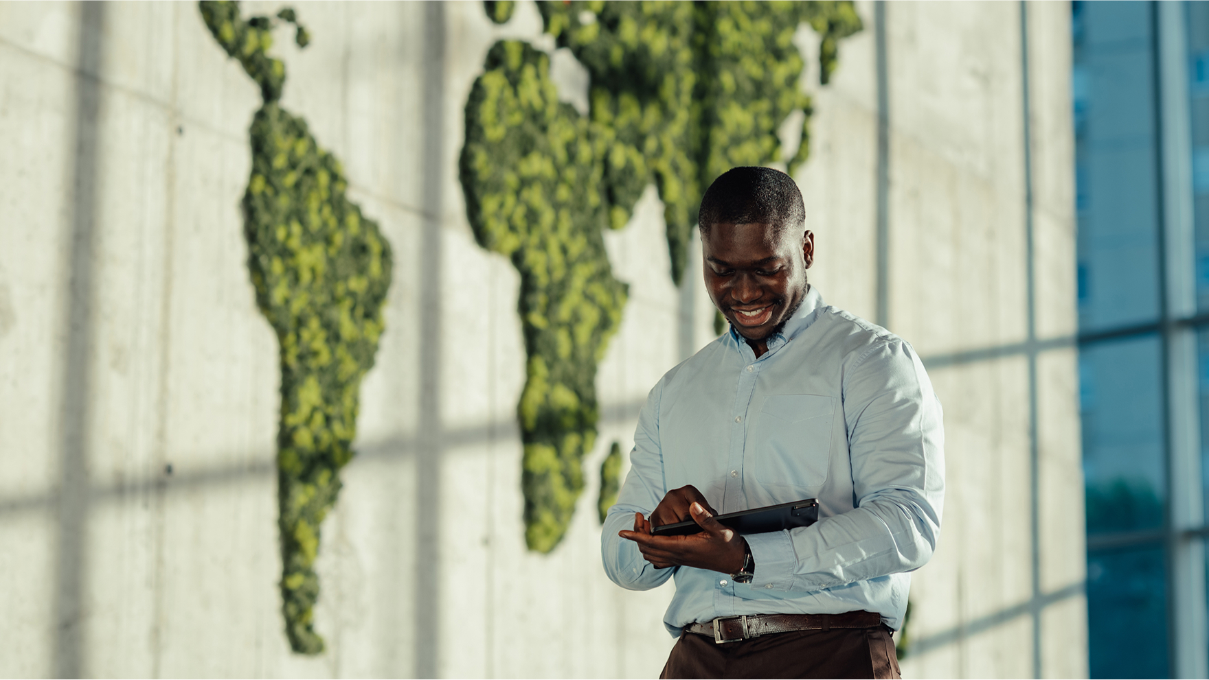A person in a light blue shirt smiles while using their phone, standing indoors in front of a green world map display on a wall.