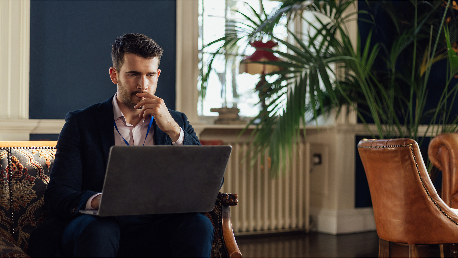 Man sitting in a stylish hotel lounge, thoughtfully working on his laptop.