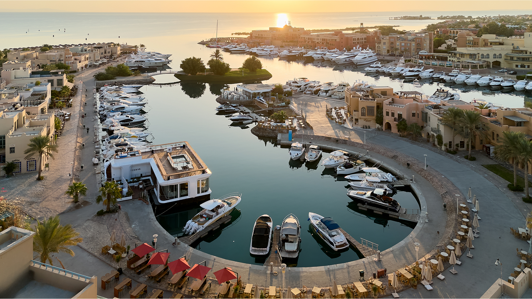 Elevated view of a harbor filled with boats, surrounded by waterfront buildings under a sunset sky.