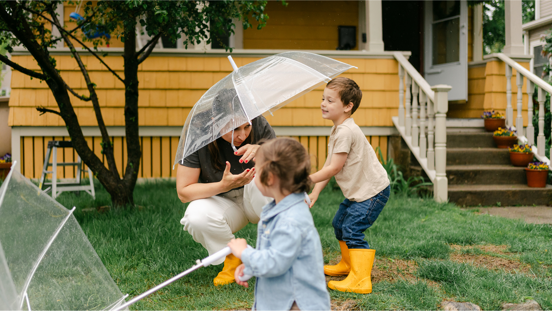 Parent and two young children playing in rain boots with umbrellas on a lawn in front of a yellow house