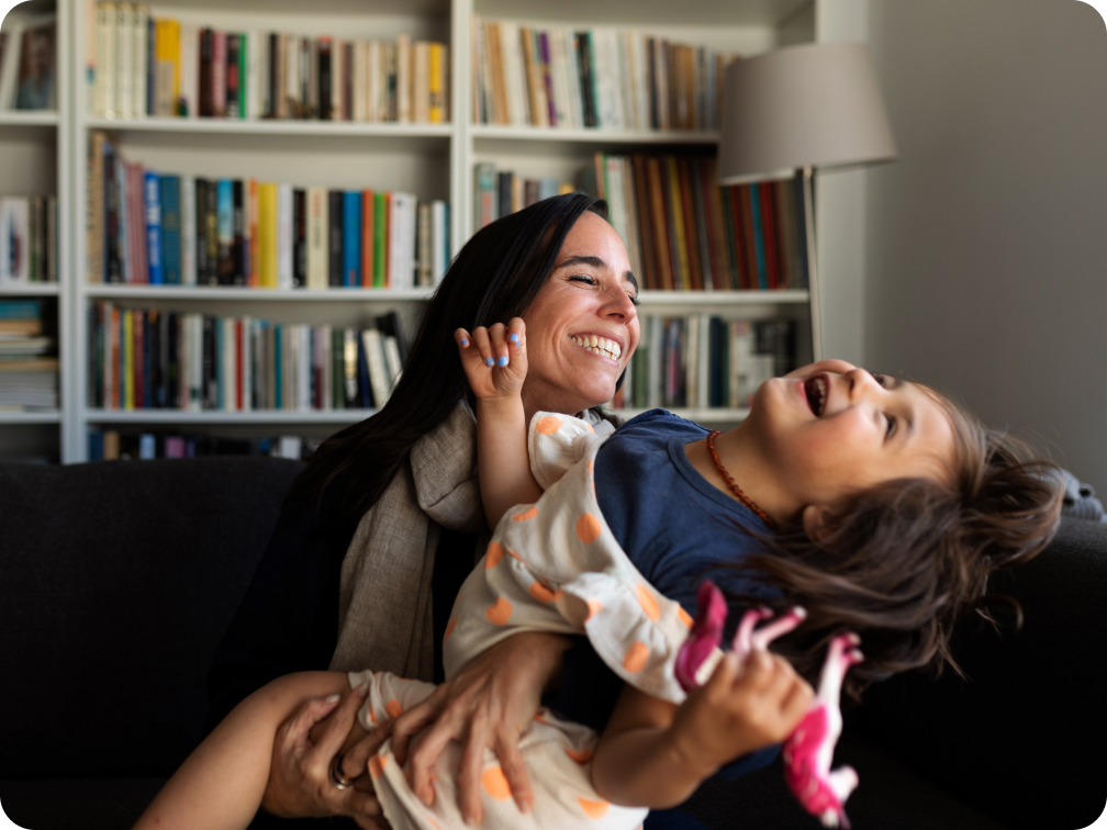 A joyful moment between a woman in a black headscarf and a young girl, both sitting on a couch in a living room lined with bookshelves. The woman lifts the girl in the air as they laugh together, surrounded by soft natural light and a stack of colorful books, highlighting their close bond and playful interaction.