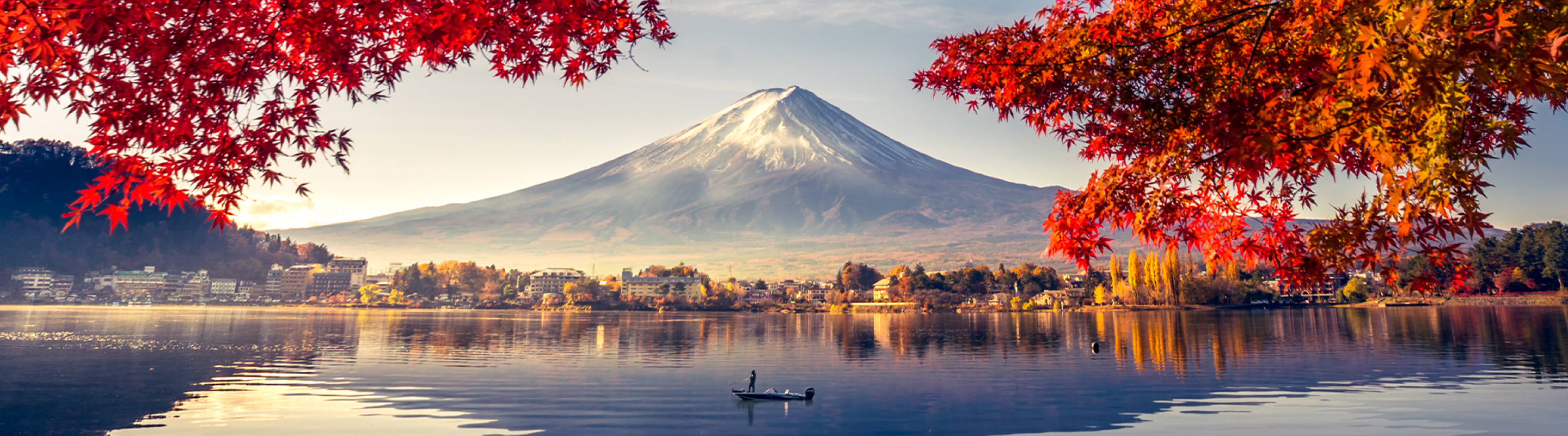 Snow-capped Mount Fuji rises beyond a calm lake, framed by vivid red maple leaves in the foreground, with shoreline buildings and a small boat reflected in the water below.