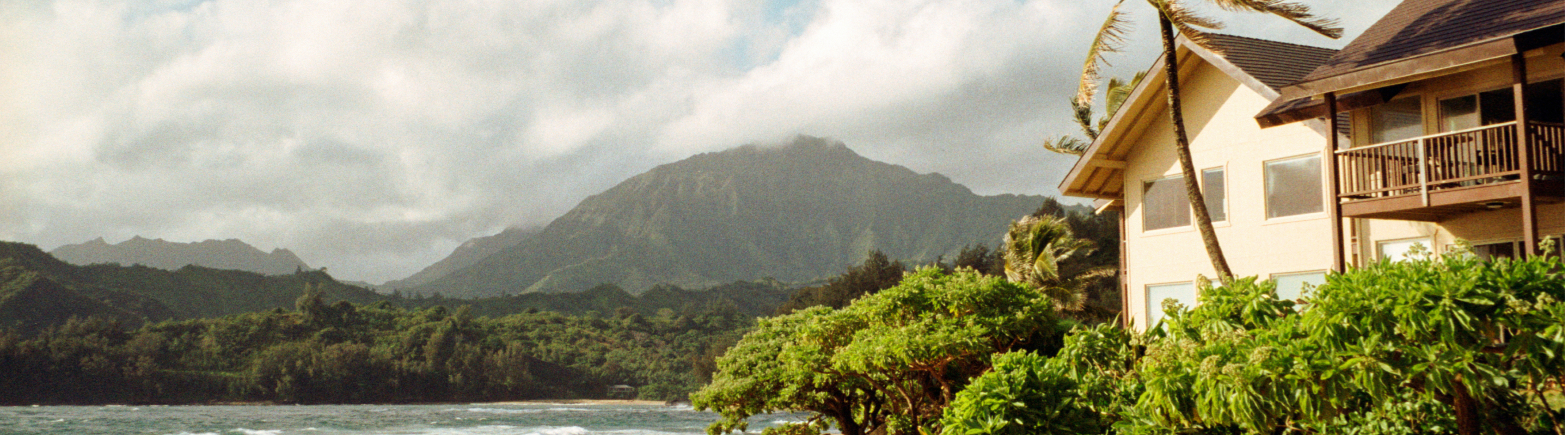 A vacation home sits beside lush greenery along a coastline, with a large mountain rising in the background under a cloudy sky.