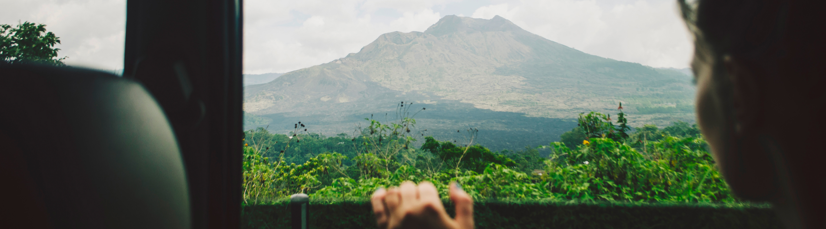 Traveler looking out a car window at a lush valley and distant mountain under a cloudy sky.