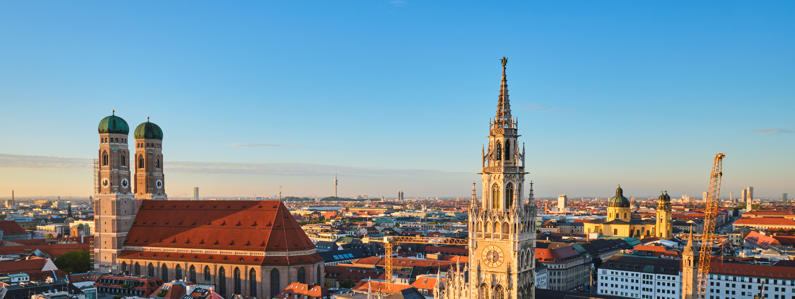 Panoramic view of Munich at sunset, featuring the Frauenkirche with its twin green domes on the left and the ornate New Town Hall (Neues Rathaus) clock tower on the right, overlooking the city’s red rooftops.